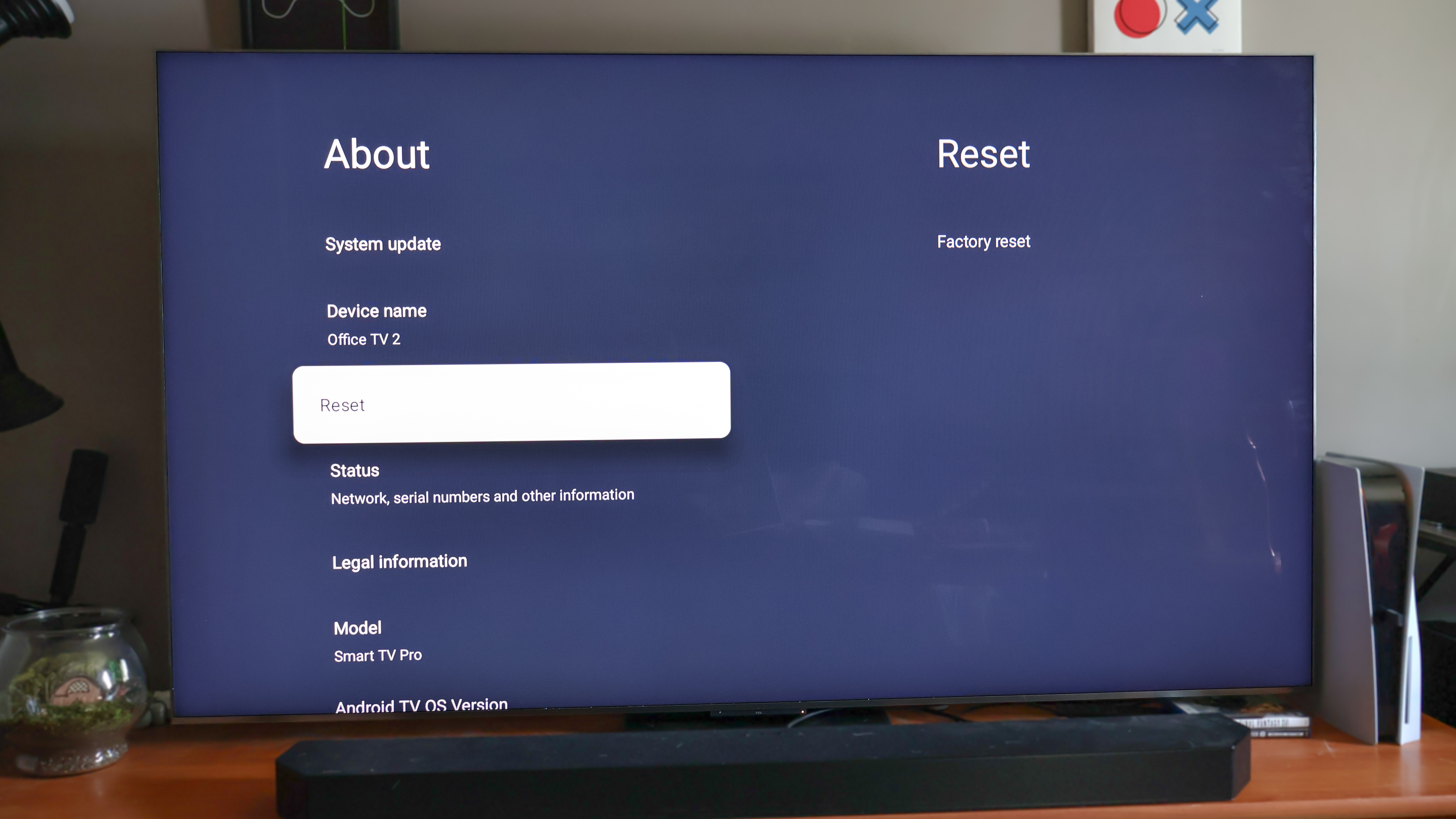 A TV on a media console displaying Google TV's System settings menu with the Factory Reset option highlighted
