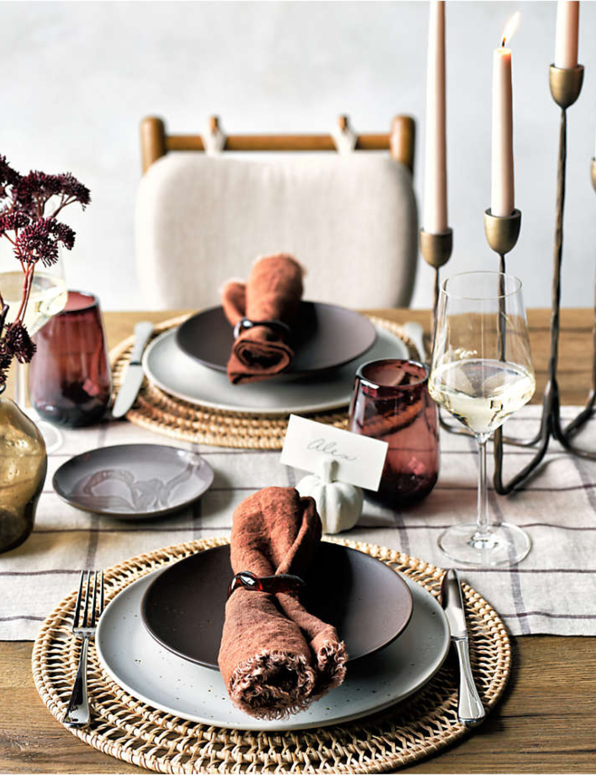 Crate & Barrel fall 2025 tableware, including woven chargers, charcoal plates, a brass candelabra, and stemware, styled on a wooden dining table.