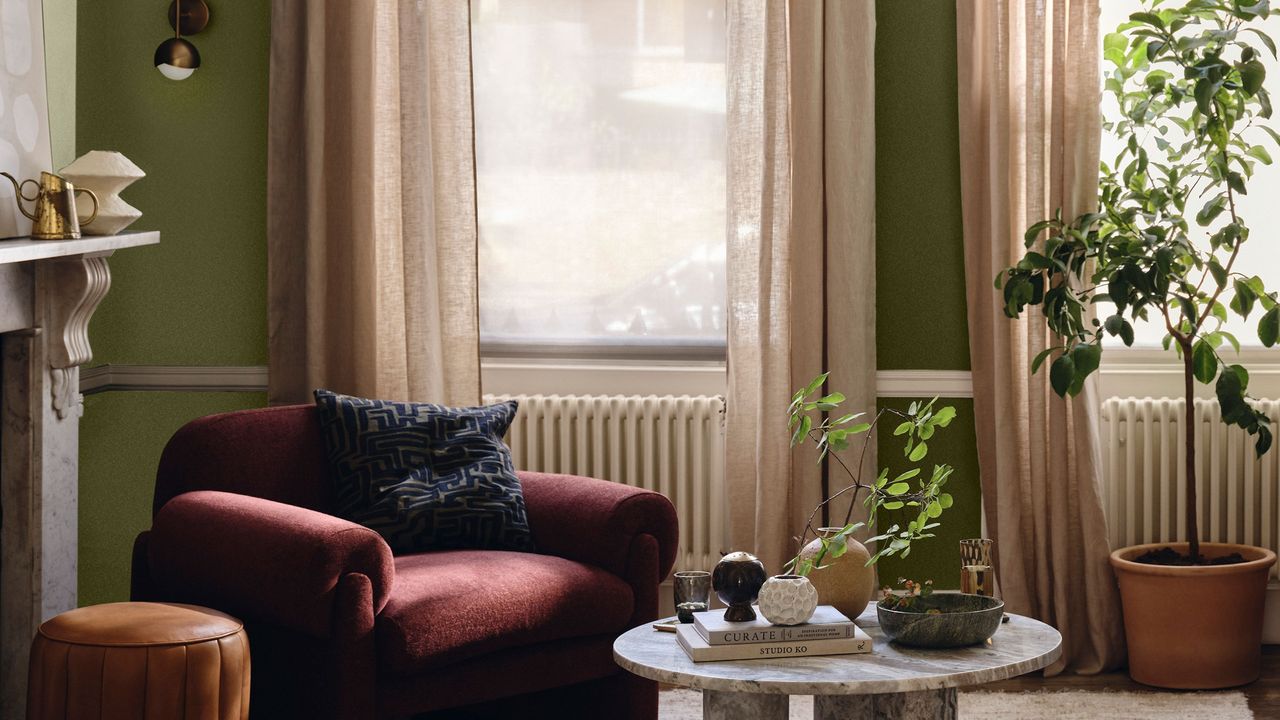 A living room painted in olive green with beige curtains and a burgundy armchair