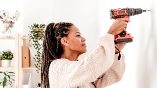 Women using orange cordless drill to drill hole into white wall of living room with shelves and plants in background