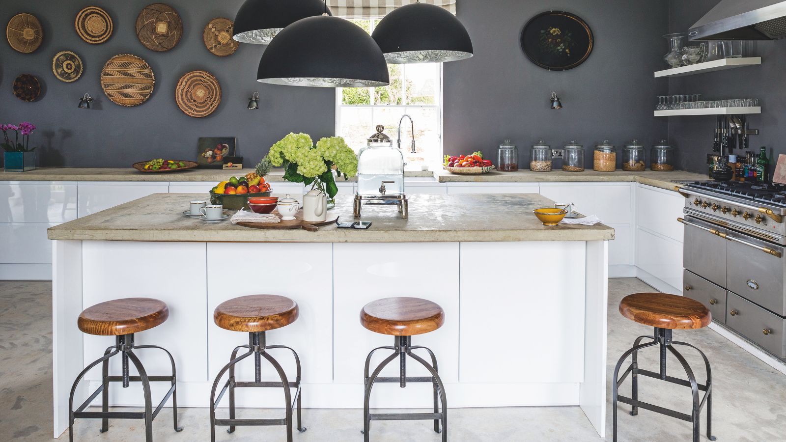 A grey kitchen with a white kitchen island with marble countertop, wooden and metal bar stools, round statement pendant lamps and decorative plates on the wall