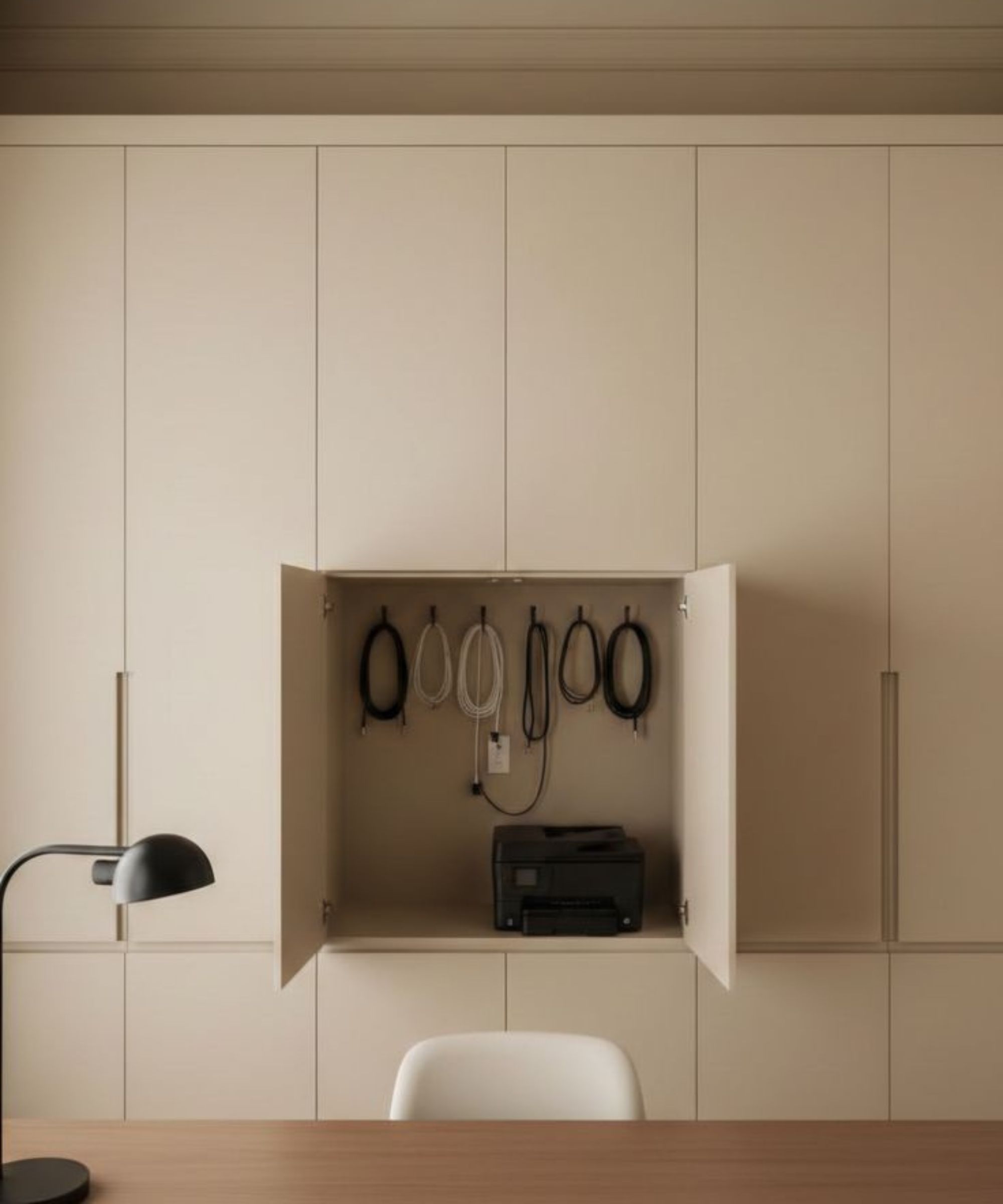 White cupboards in study, opening showing hanging wires inside, dark wood desk with lamp placed in front