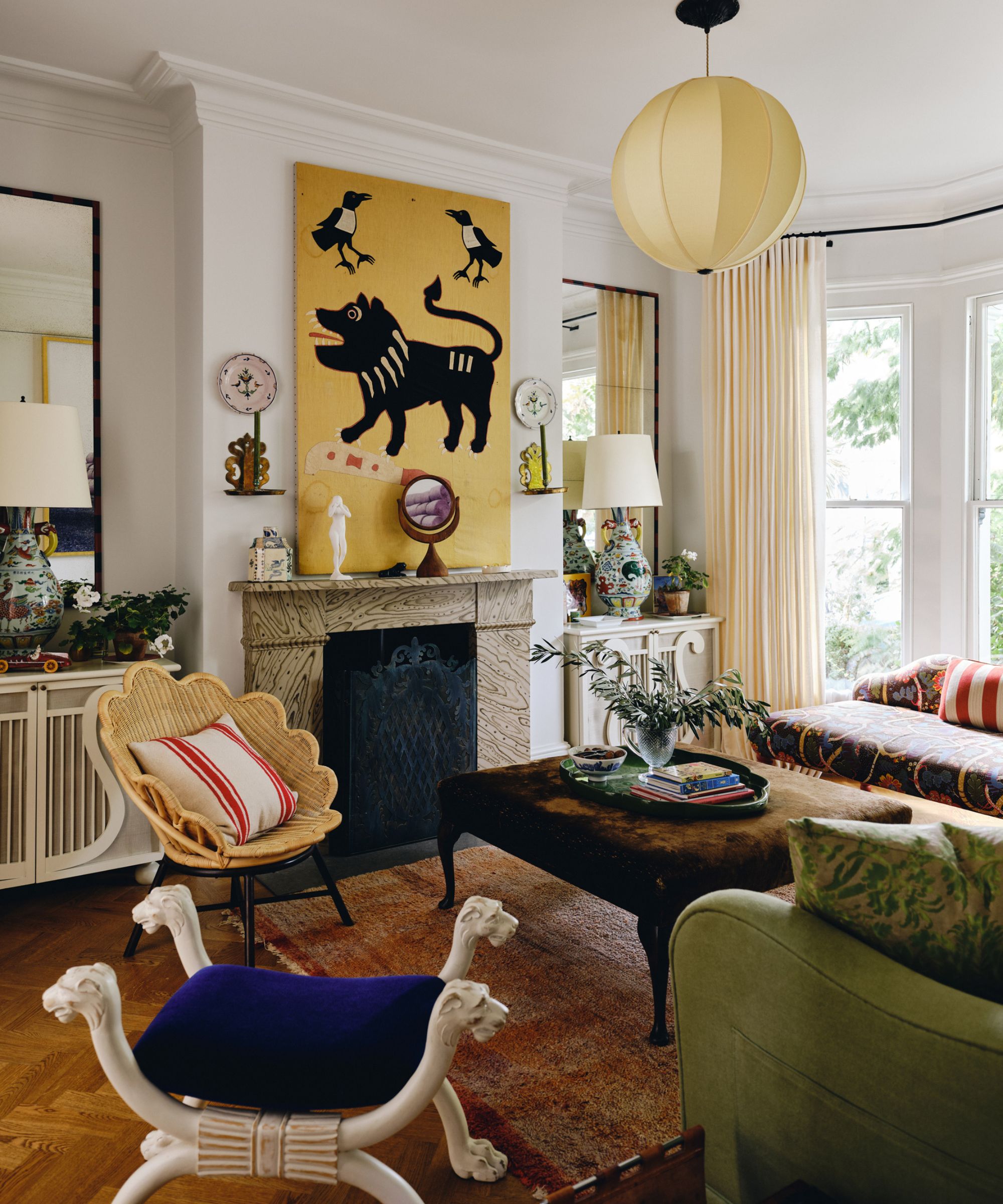 image of an eclectic living room with white walls and a large yellow painting above a marble fireplace with a scalloped rattan chair and eclectic furniture