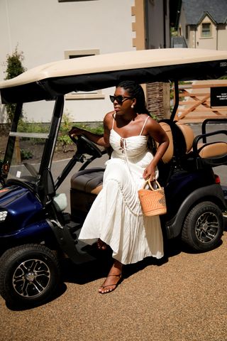 Remy sits in gold cart wearing white summer dress, basket bag and sandals