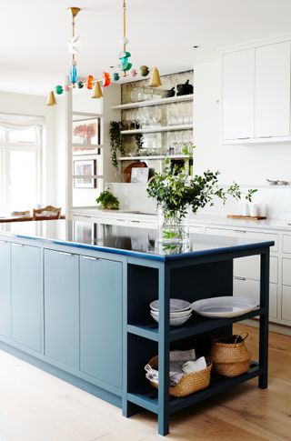 An all white kitchen with hardwood floors and a dark blue kitchen island. The island has a lava stone worktop and there is a clear vase with green stems on the counter. A colorful light fixture hangs above the island.