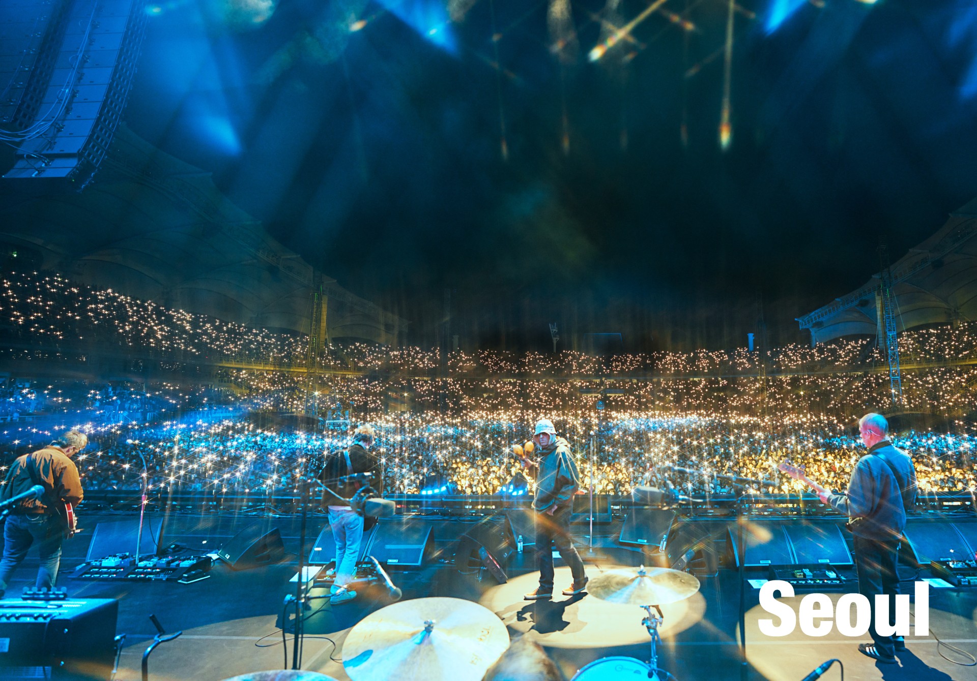 A wide, high-angle color shot shows the band performing on a massive stage in Seoul, facing a stadium filled with thousands of glowing phone lights.