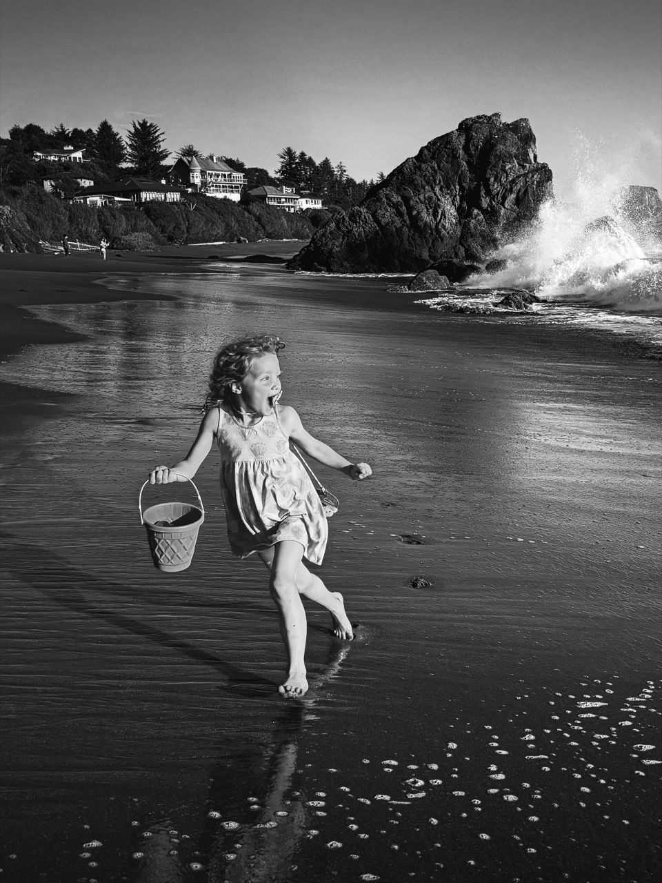 Photograph of a young girl on the beach, holding a bucket and running away from a crashing wave, in black &amp; white