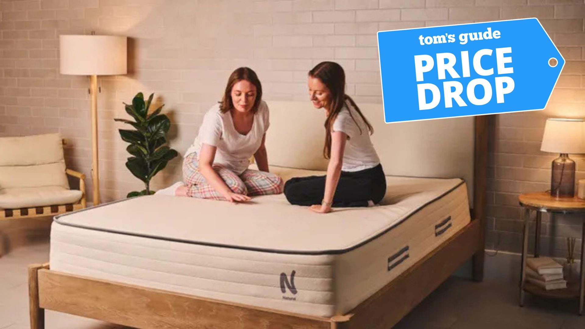Two Tom's Guide mattress testers sit on the Nolah Natural 11 mattress. A deals badge is seen in the top right foreground. 