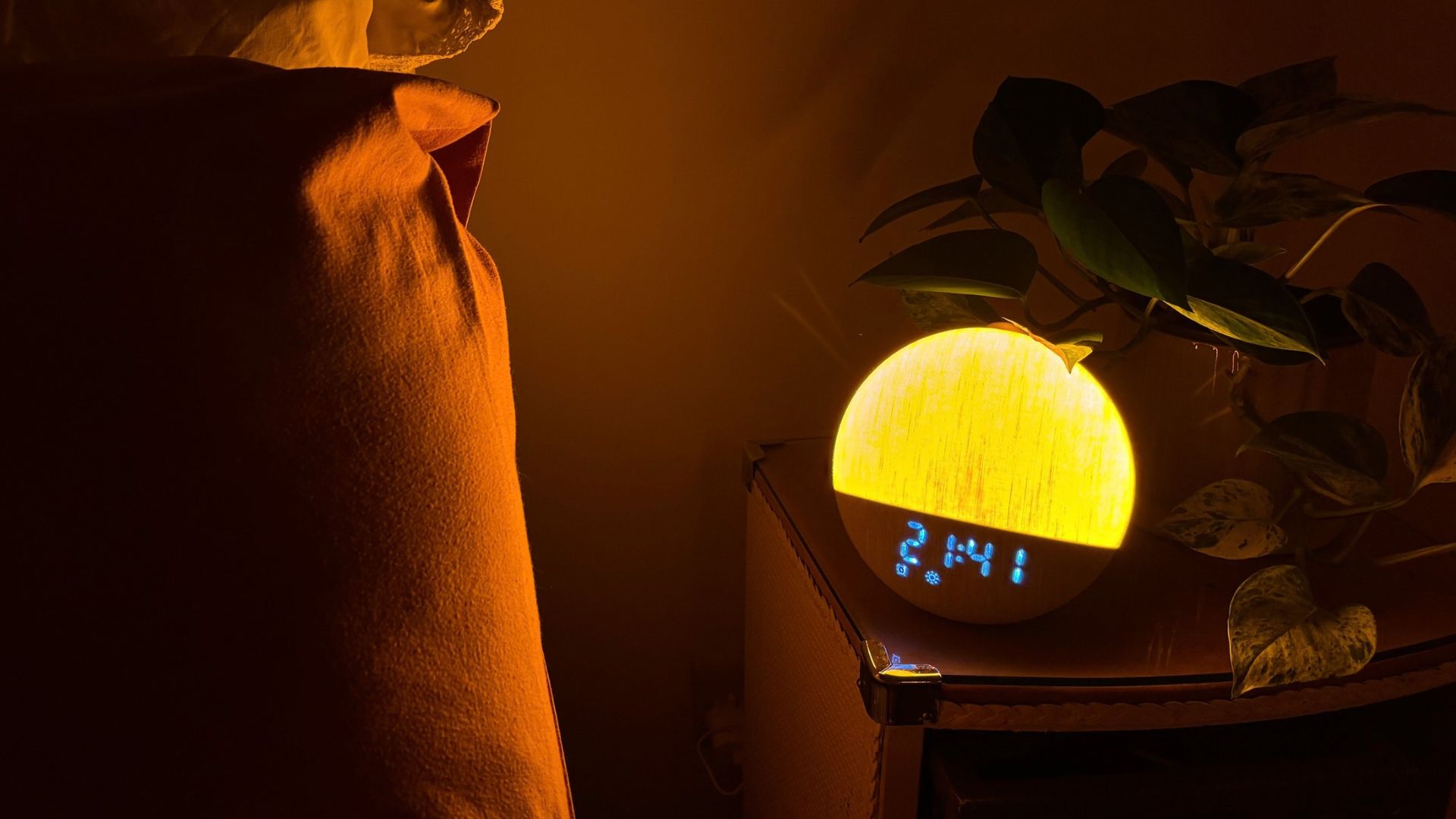 The circular Dreamegg Sunrise 2 sunrise alarm clocks glows orange as it sits on a nightstand in an otherwise dark bedroom, placed next to a green pot plant. The display time read: 21:41