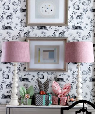 Hallway console style with two table lamps, wrapped gifts, bowl of small easter eggs, black and white rabbit wallpaper, artwork on walls