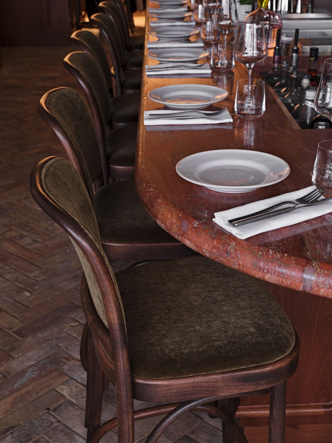 A red marble bar counter is decorated with sleek tableware, velvet green and wooden stools, and herringbone floors.