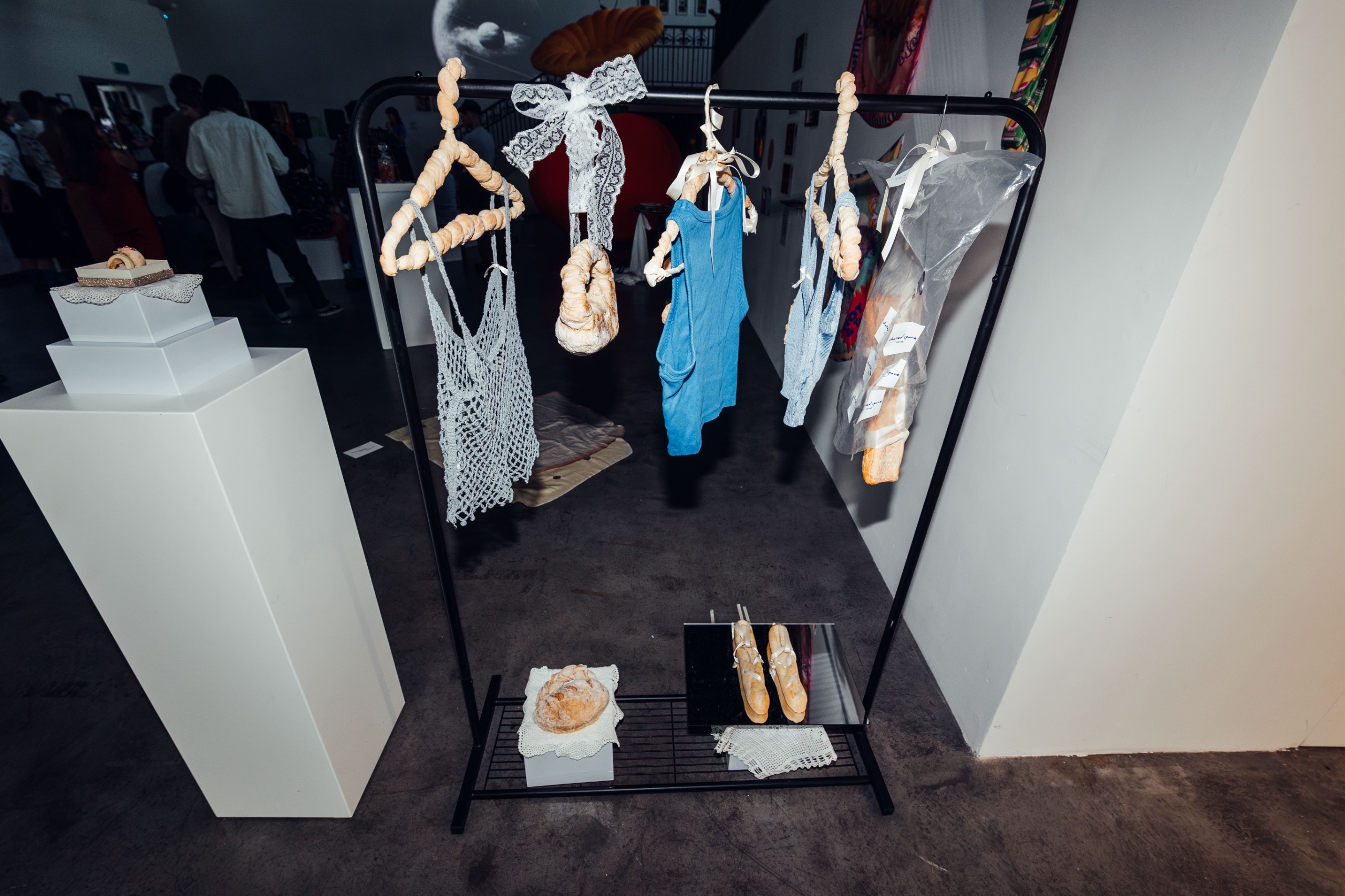 A food and fashion installation with clothes hanging on bread-made hangers in a dimly lit gallery with dark floors and white walls.