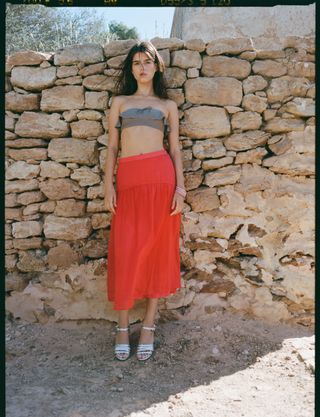 Model wearing a taupe strapless bikini top with a red A-line drop-waist skirt and silver heels from the J.Crew x Maryam Nassir Zadeh collection.