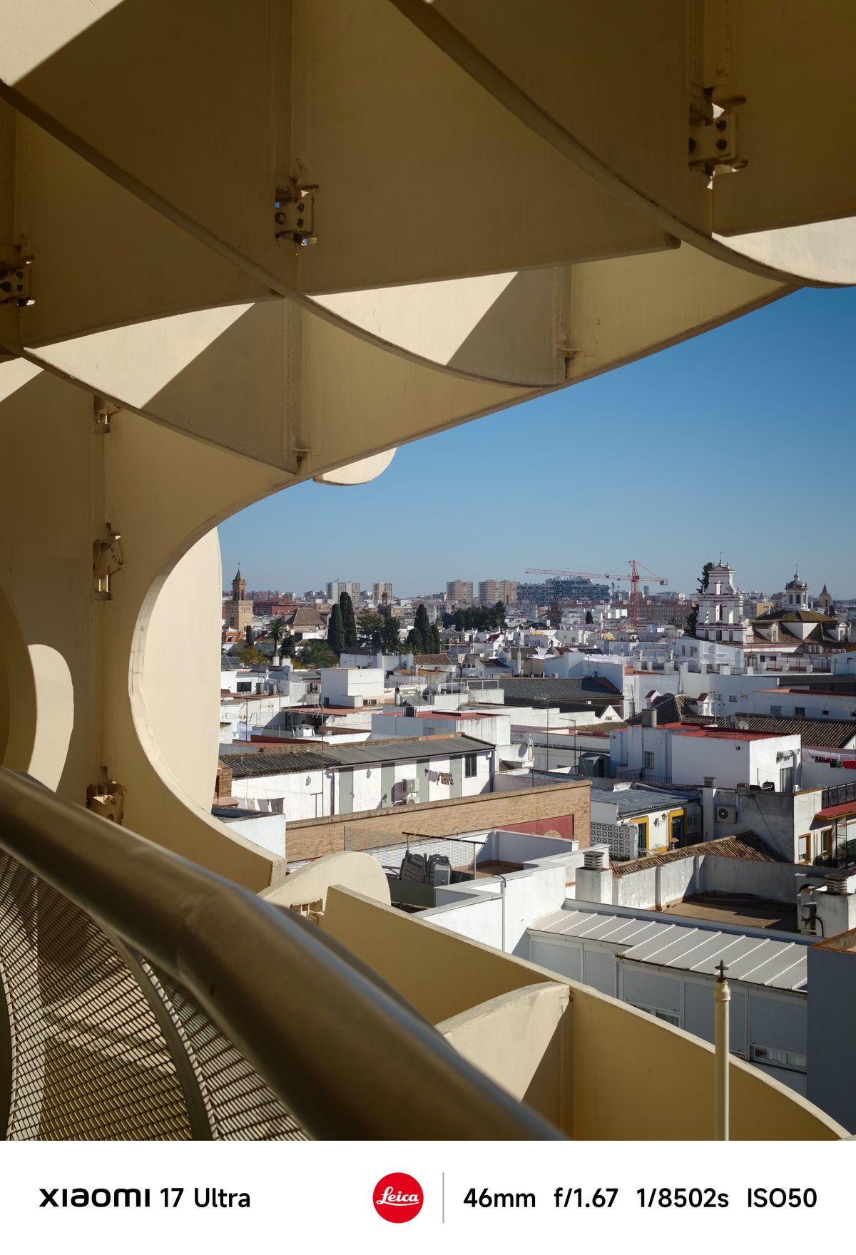 Cityscape of white rooftops framed by the Parasol&rsquo;s curved supports against a blue sky.