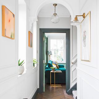 a hallway with white panelled walls and wooden herringbone flooring facing into a living space with green velvet sofa