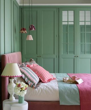 A green bedroom with a pink bed and a white nightstand with a table lamp