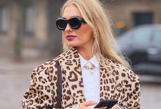 A woman wearing oversize black sunglasses and a gold necklace with a cheetah-print coat layered over a white button-down.