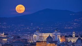 a large slightly orange moon rises behind thin wispy clouds with a mountain visible to the right, in the foreground is a large city with many old buildings.