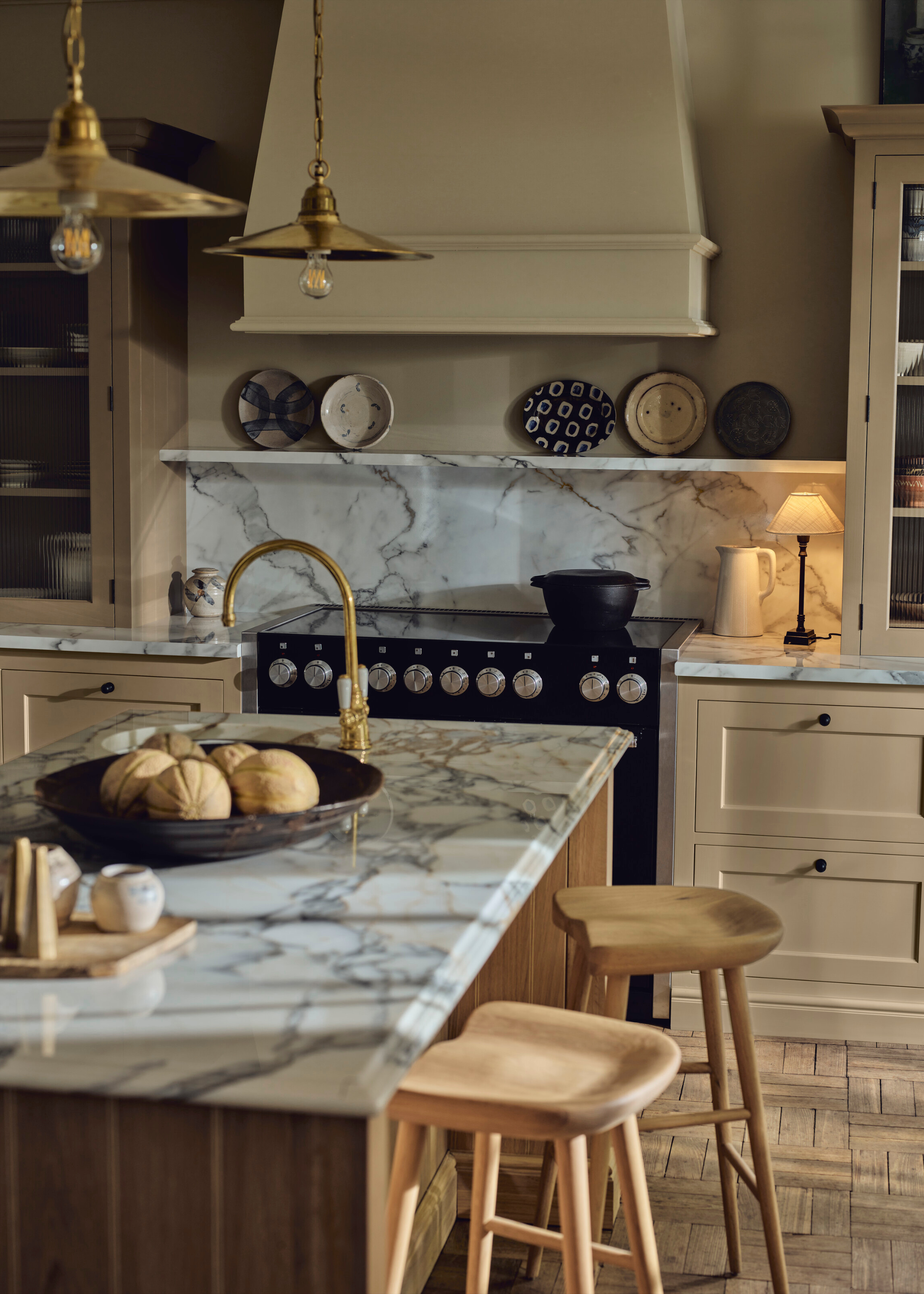 A creamy yellow, neutral painted kitchen with white marble worktops, wooden bar stools at the island and brass lighting hung over and brass tap for the island sink. A kitchen lamp sets the ambiance well in a darker corner.