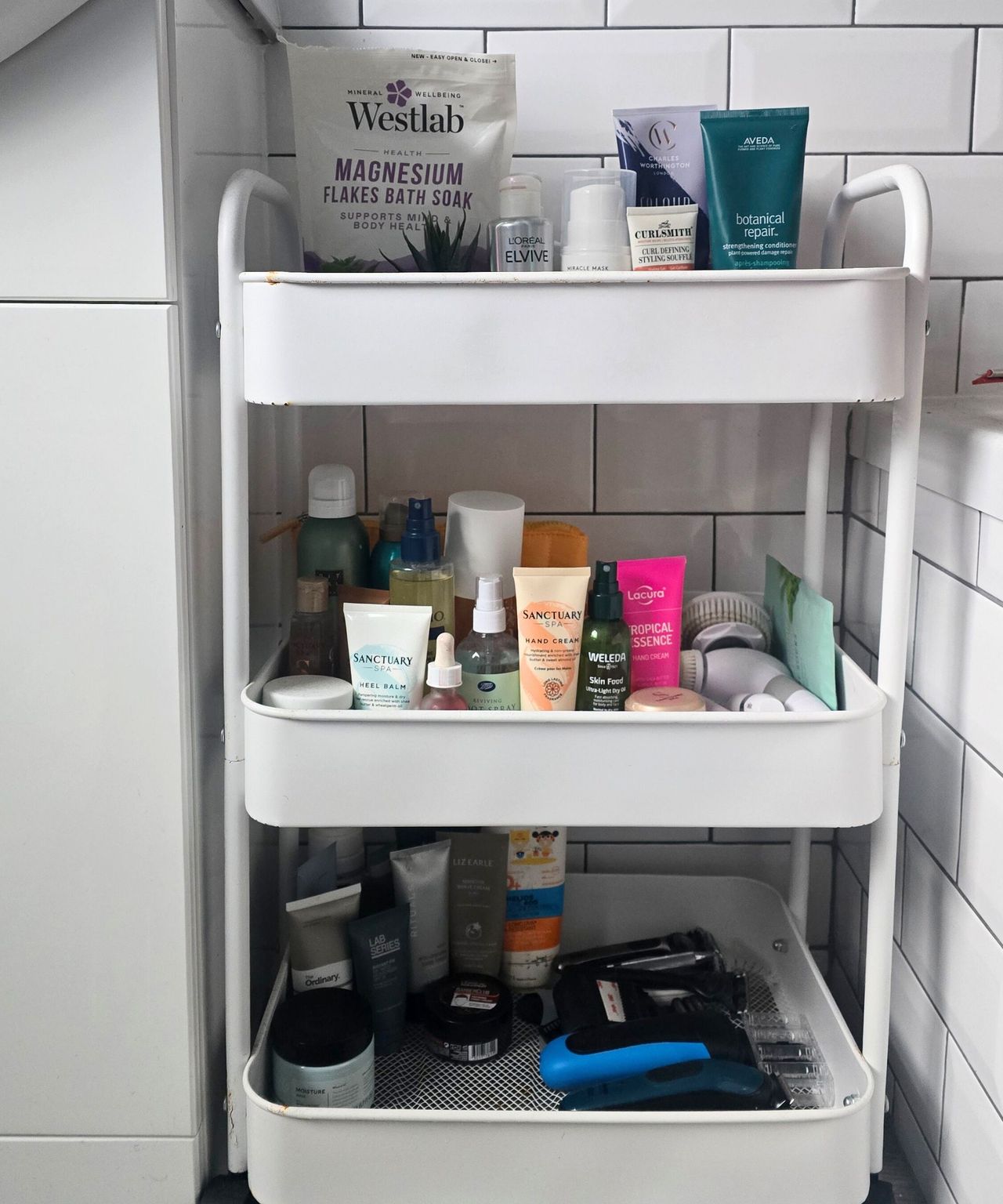 A tidy three-tier white storage trolley with colorful toiletries on top of it and white tiles behind it