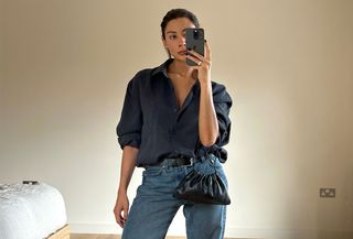 A woman standing in a bedroom with a beige wall taking a mirror selfie wearing a navy blue button-down top and jeans