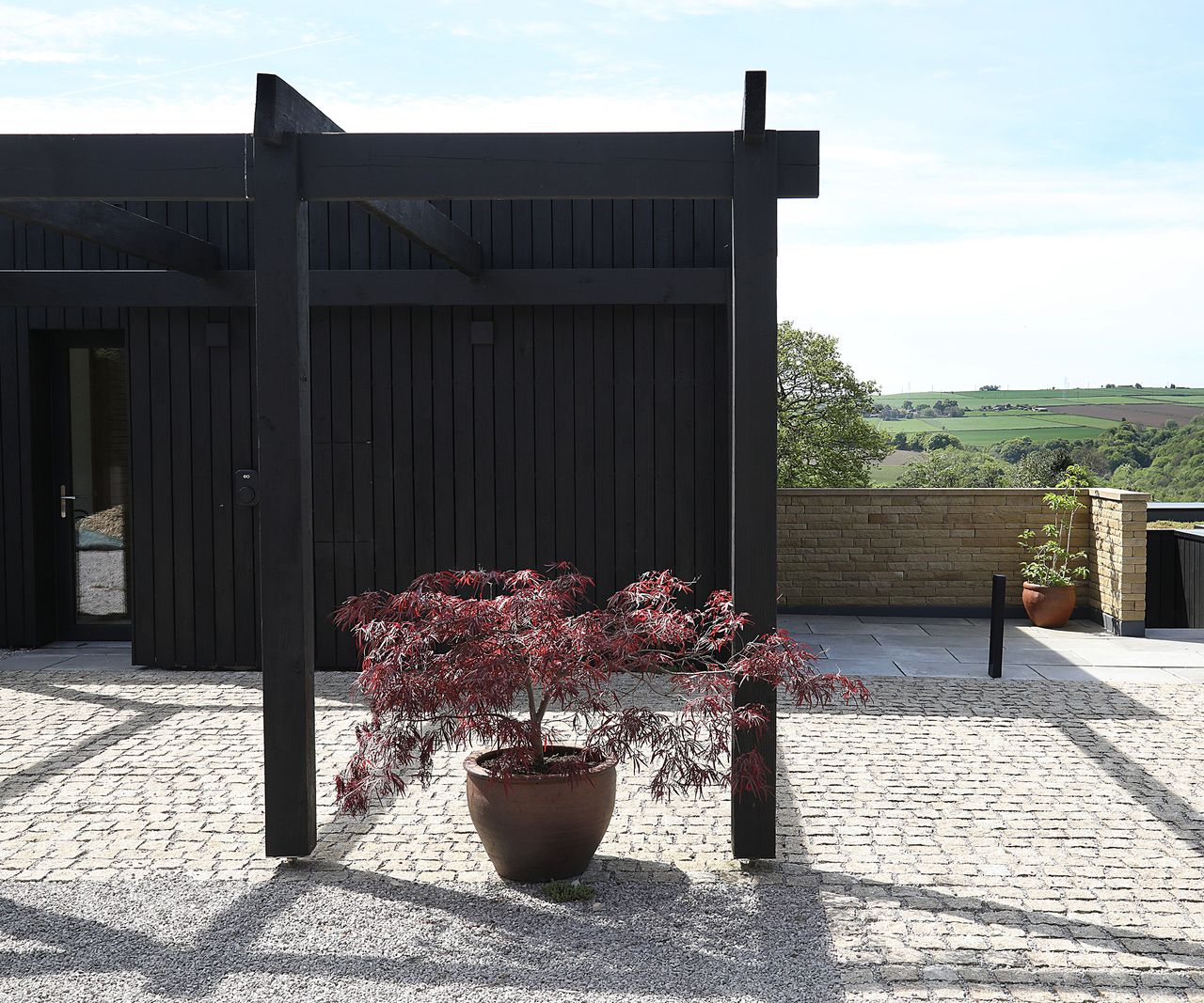 A single ornamental acer tree sits in a clay pot on brick paved driveway. The home itself is a black clad timber newly constructed self build home