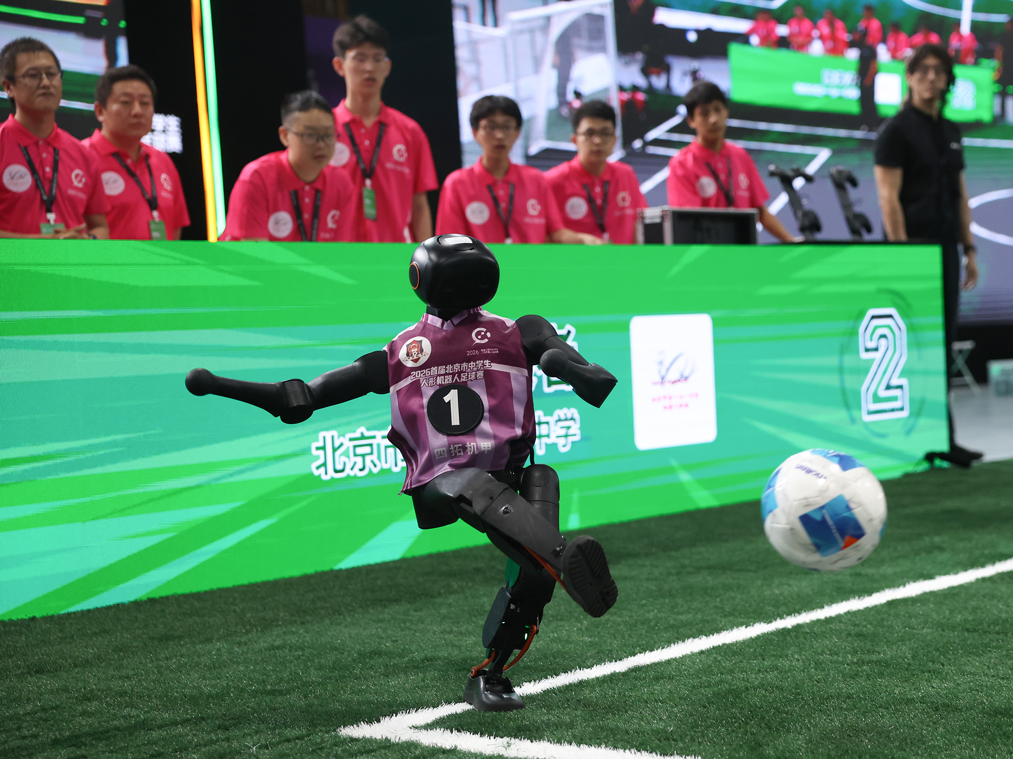 A robot in a football jersey kicks a white ball as people behind watch.