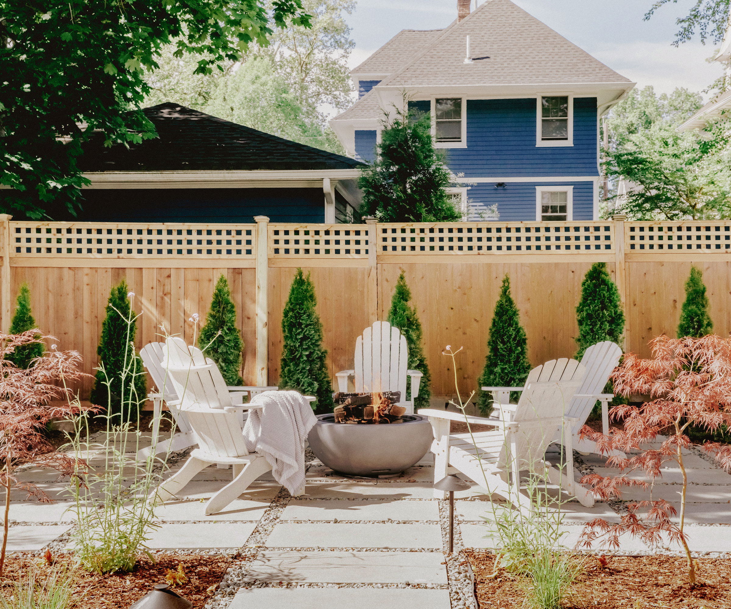 fire pit with garden furniture set up around it including Adirondack chairs, clipped evergreens, a fence topped with trellis, pavers and gravel