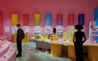 Three people, two women and one man, stand in a pink-shaded museum showcasing Barbie dolls and artifacts.