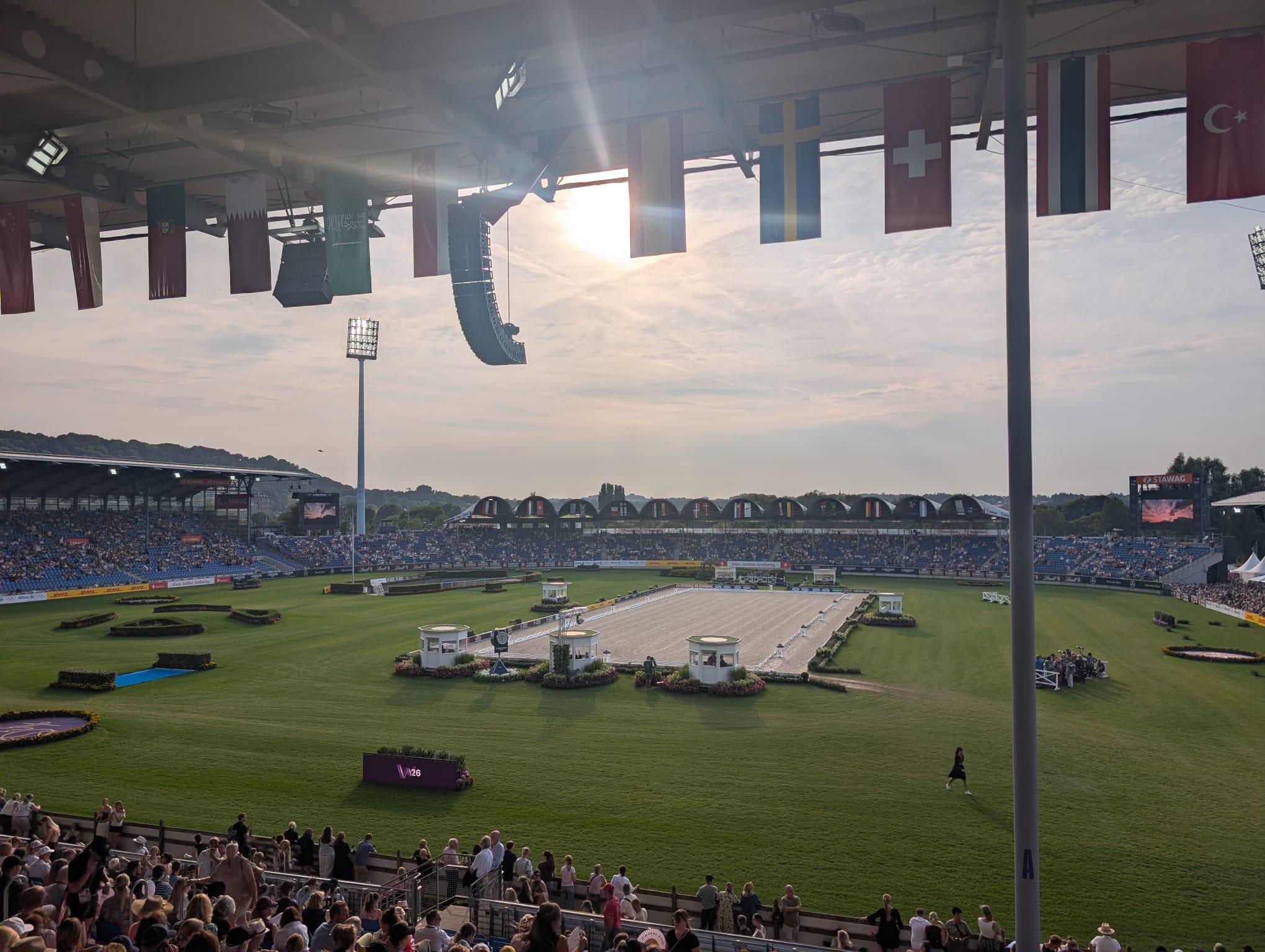 Image shows Aachen's main arena in the evening light &amp;ndash; in the distance is a sand dressage arena surrounded by grass, in the foreground are full stands