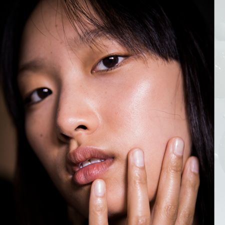 A woman with glowing skin with four of her fingers resting against her cheek. She is staring into the camera.