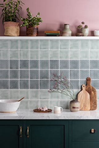 A blue kitchen with blue tiled backsplash and pink wall paint decor, floating white shelf with houseplants, marble worktop with chopping boards and navy blue cabinets