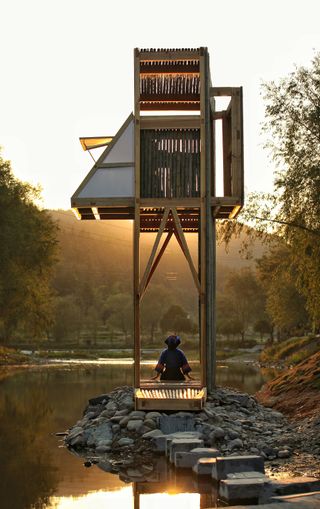 A shelter that looks like a tree house on the ground is set next to a river, with a woman sitting on the rocks below it.