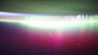 A streak of white showing Comet Lemmon moves into a red and green aurora seen on the edge of Earth in space