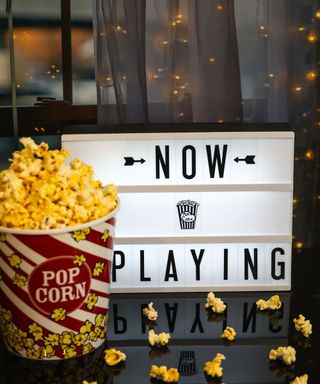 A white rectangular lightbox that says 'now playing' in black lettering, with a tub of popcorn next to it, popcorn kernels on the black table, and a gray curtain with fairy lights around it behind