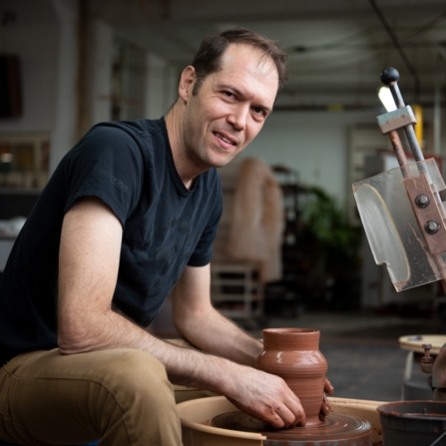 Image of a man in a black short-sleeve shirt and khakis sitting at a pottery wheel in a pottery studio