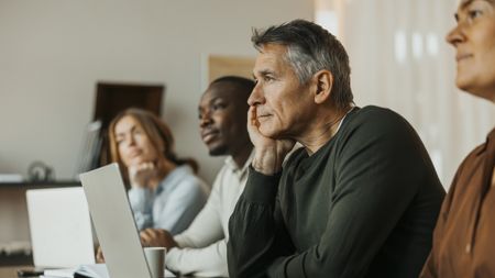 A diverse set of employees sits at a meeting table listening to a speaker off-camera.