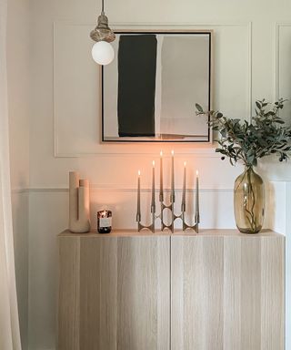 A white wall with a white and black wall art print and a wooden console table with candles and a plant on it