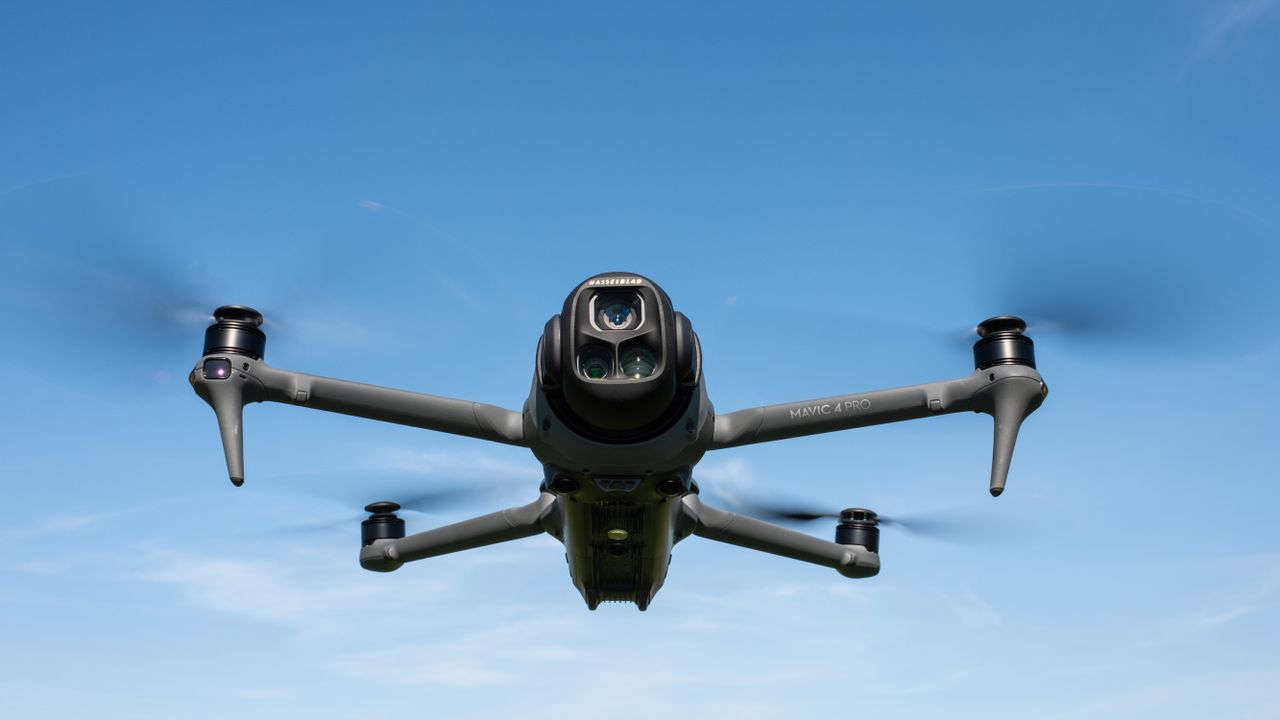 DJI Mavic 4 Pro in flight against a blue sky facing towards the camera with the drone camera and gimbal visible