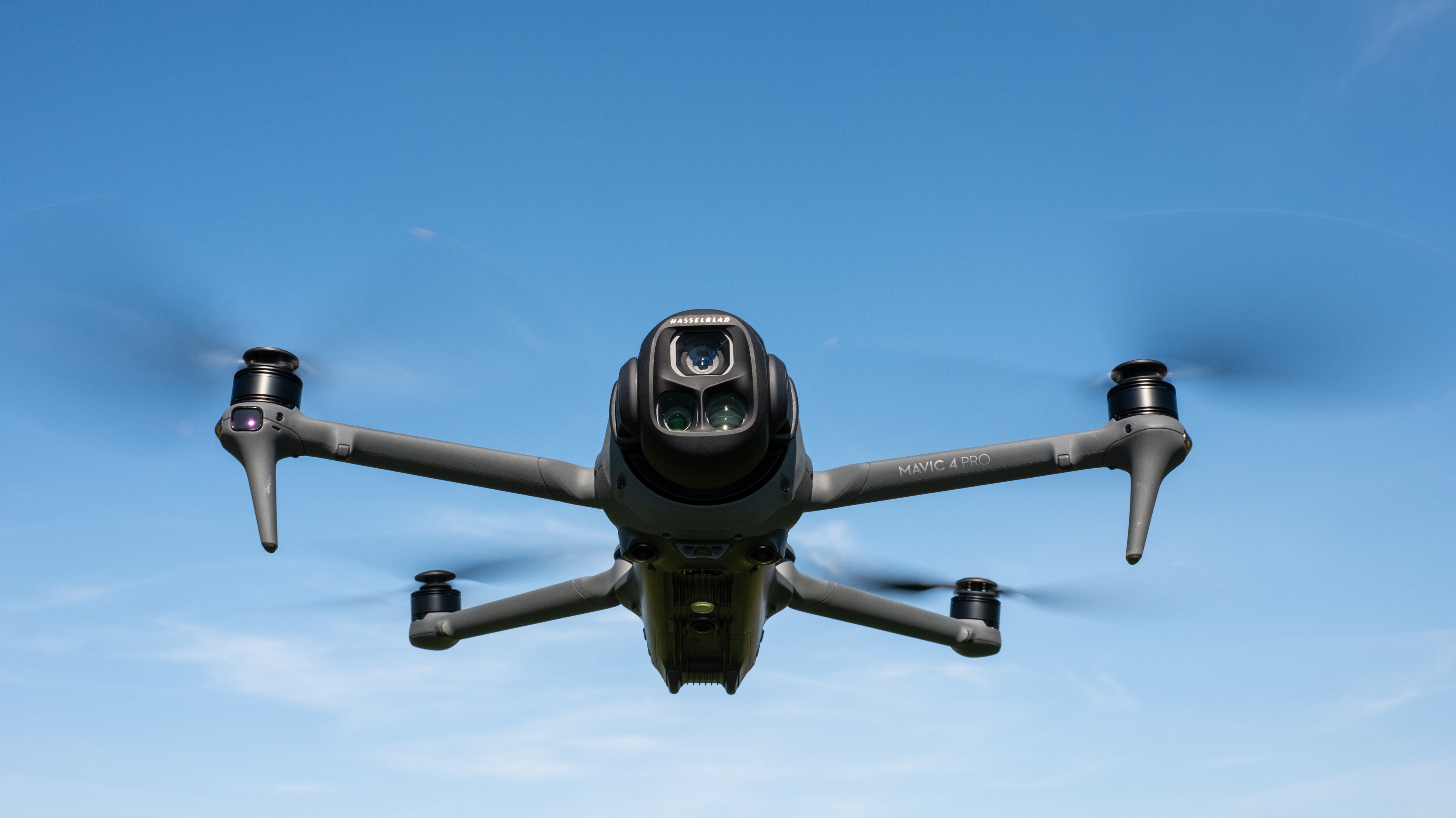 DJI Mavic 4 Pro in flight against a blue sky facing towards the camera with the drone camera and gimbal visible