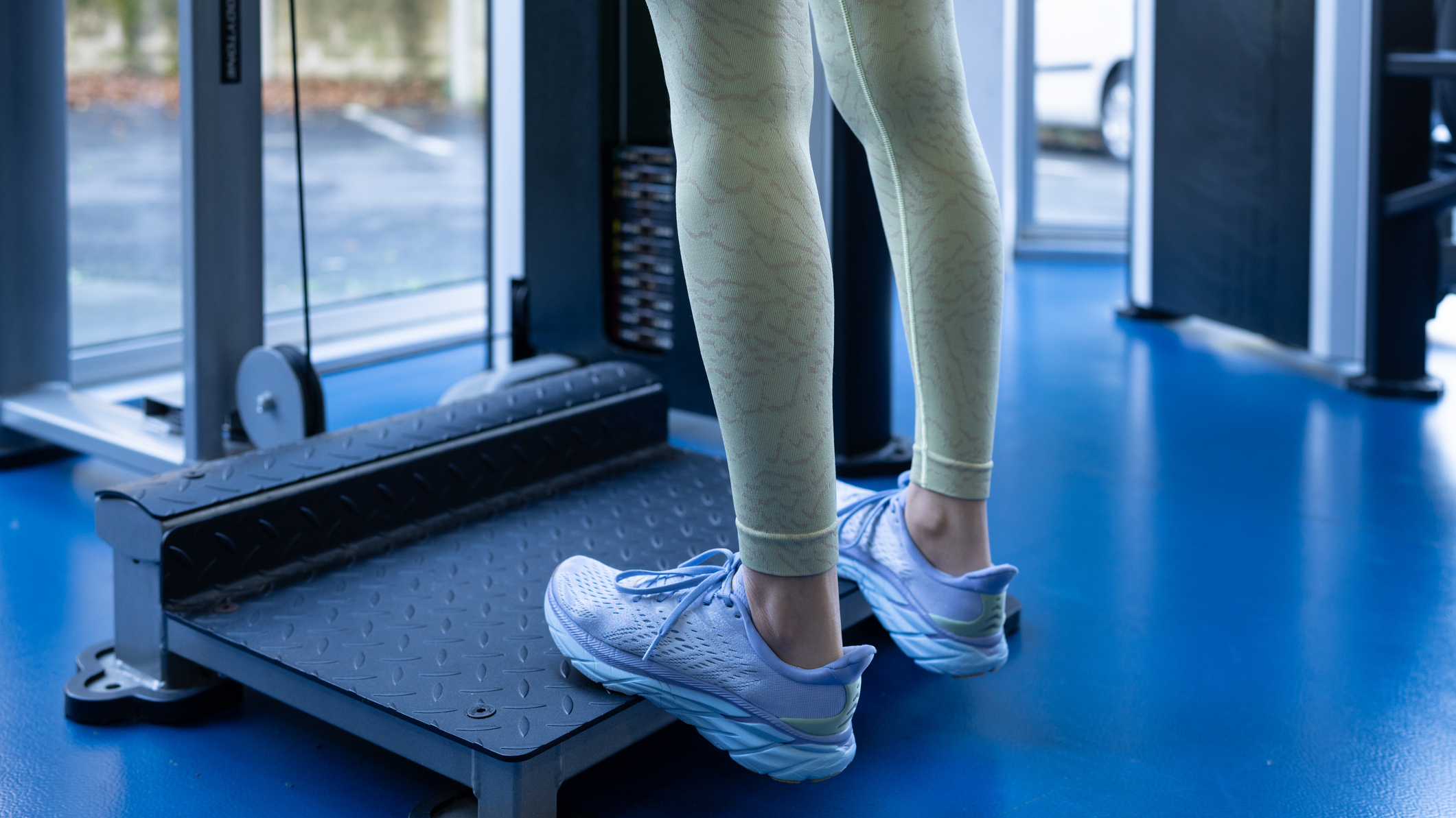 A woman in leggings and gym shoes stands on a platform with her heels hanging off the edge