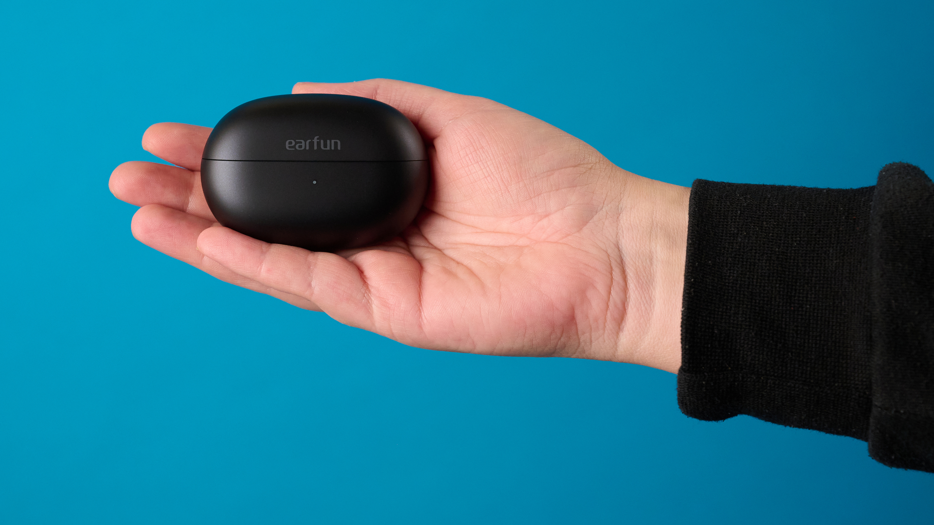A hand holding the EarFun Clip 2 in black photographed in front of a blue background
