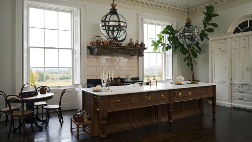 Kitchen in large room with high ceilings, large sash windows, corniced ceiling, wooden marble topped kitchen island, large pendant lighting with small round dining table and four chairs in corner 