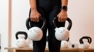 Woman holding two kettlebells in front of her thighs, two outline of breasts are drawn on each kettlebell in black marker