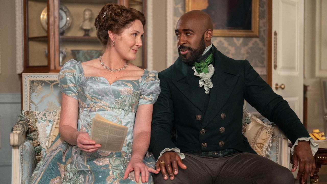 左から右へ：レディ・ヴァイオレット・ブリジャートン役のルース・ジェメルとアンダーソン卿役のダニエル・フランシス。彼らはソファに一緒に座って小指を触っています。 