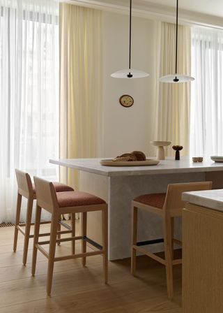 A clean kitchen space with bar stools and a large marble island/ dining space. Two pendant lights also hang above.