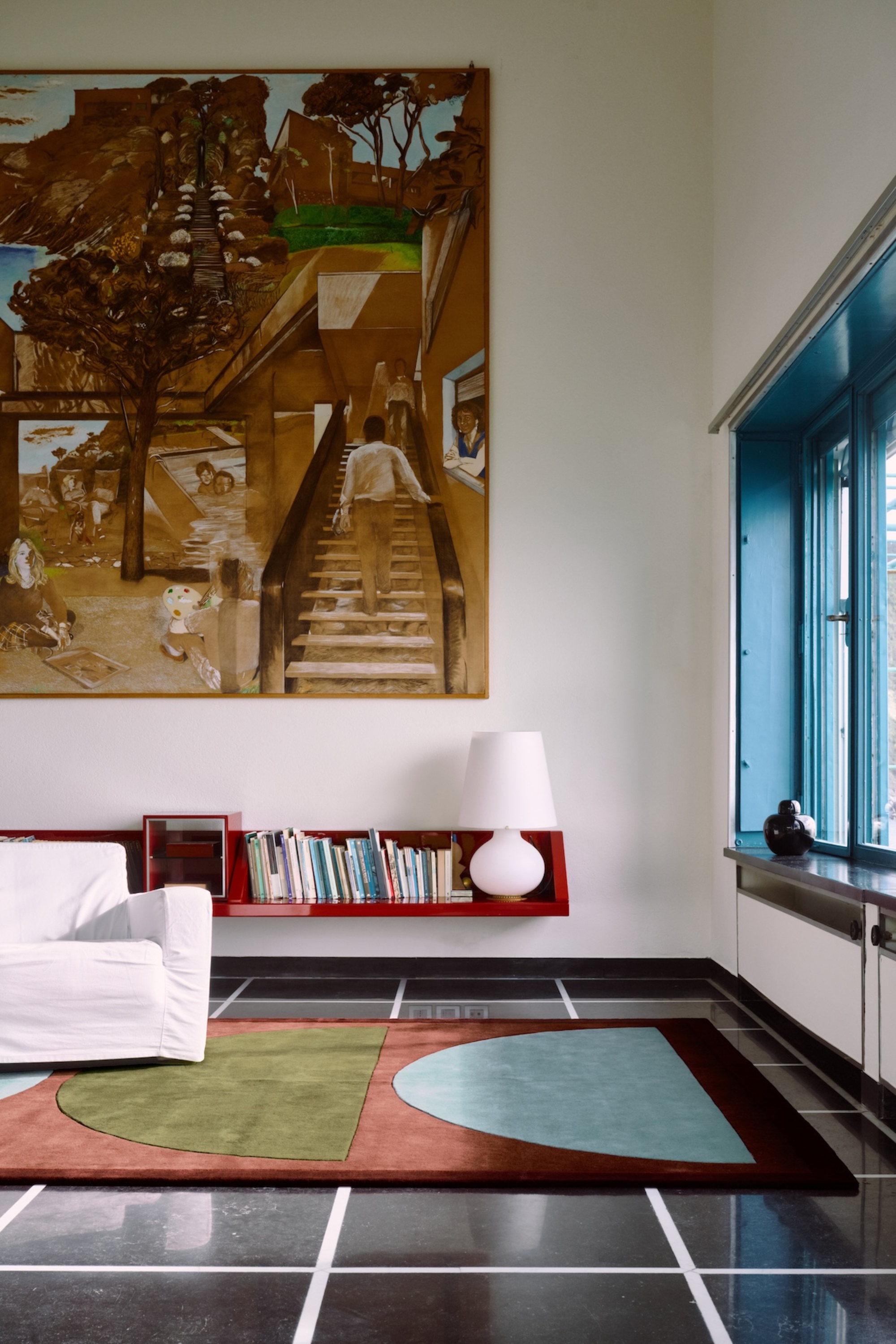modern room with white walls, large artwork, blue window frames, floating red bookshelf with table lamp, white slipcover sofa, and colorful graphic rugs on a black glossy floors