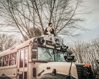 caleb on top of his converted school bus with his dog, with trees in the background