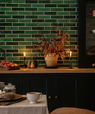 A kitchen with dark green tiles, a wooden worktop with apples and autumn leaves in a vase and a white table with hot chocolate on it