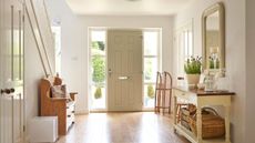 Hallway ideas are so useful. Here is a white hallway with a beige door with glass panels either side, a light wooden bench next to a staircase and a white console table with a plant on it and crate underneath it